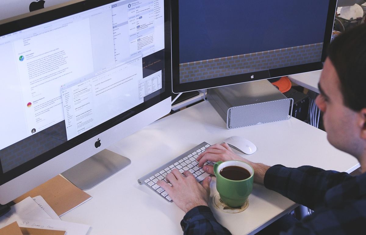 Personne assise à un bureau devant deux moniteurs d'ordinateur et un clavier, tenant un café Personne assise à un bureau devant deux moniteurs d'ordinateur et un clavier, tenant un café